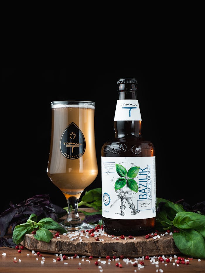 A craft beer bottle and glass on wood with basil leaves and pepper around, set against a dark background.